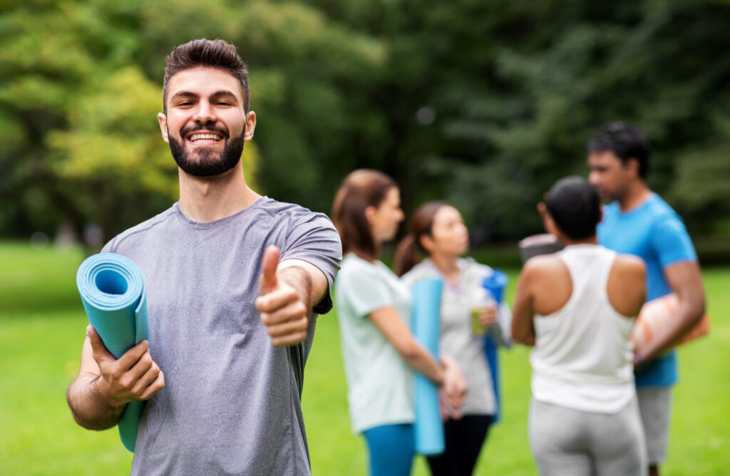 Smiling man holding a yoga mat and giving a thumbs-up in a park with a group of coworkers preparing for a wellness activity in the background, representing employee wellness and teamwork.