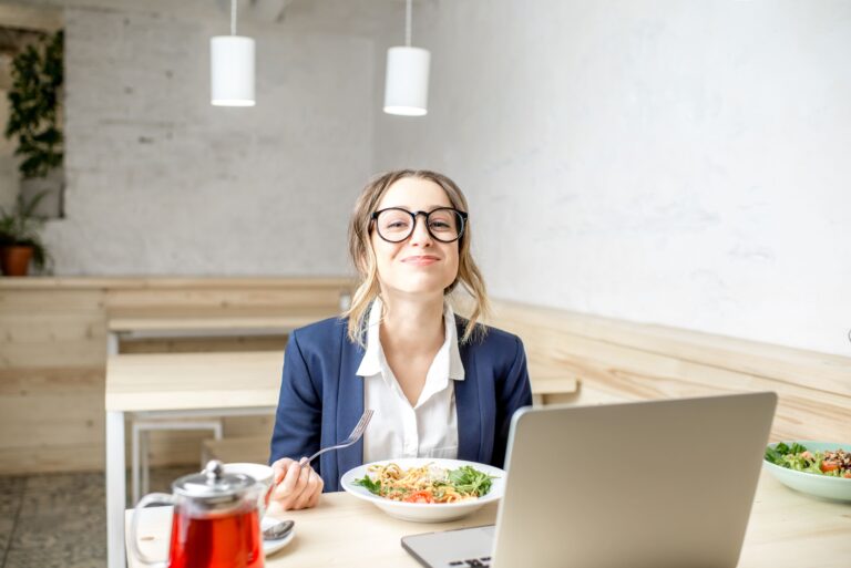 Smiling businesswoman enjoying a healthy lunch at her desk while working on a laptop in a bright office café.