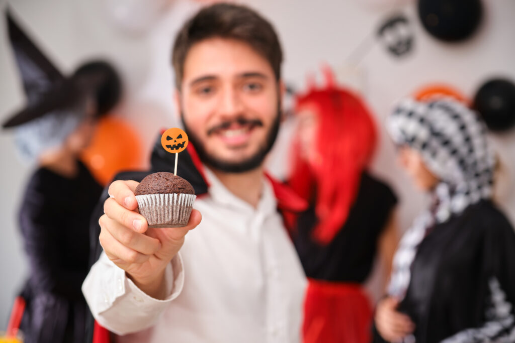 Young man in a Halloween costume holding a festive cupcake with a pumpkin topper, with coworkers in costumes celebrating in the background.