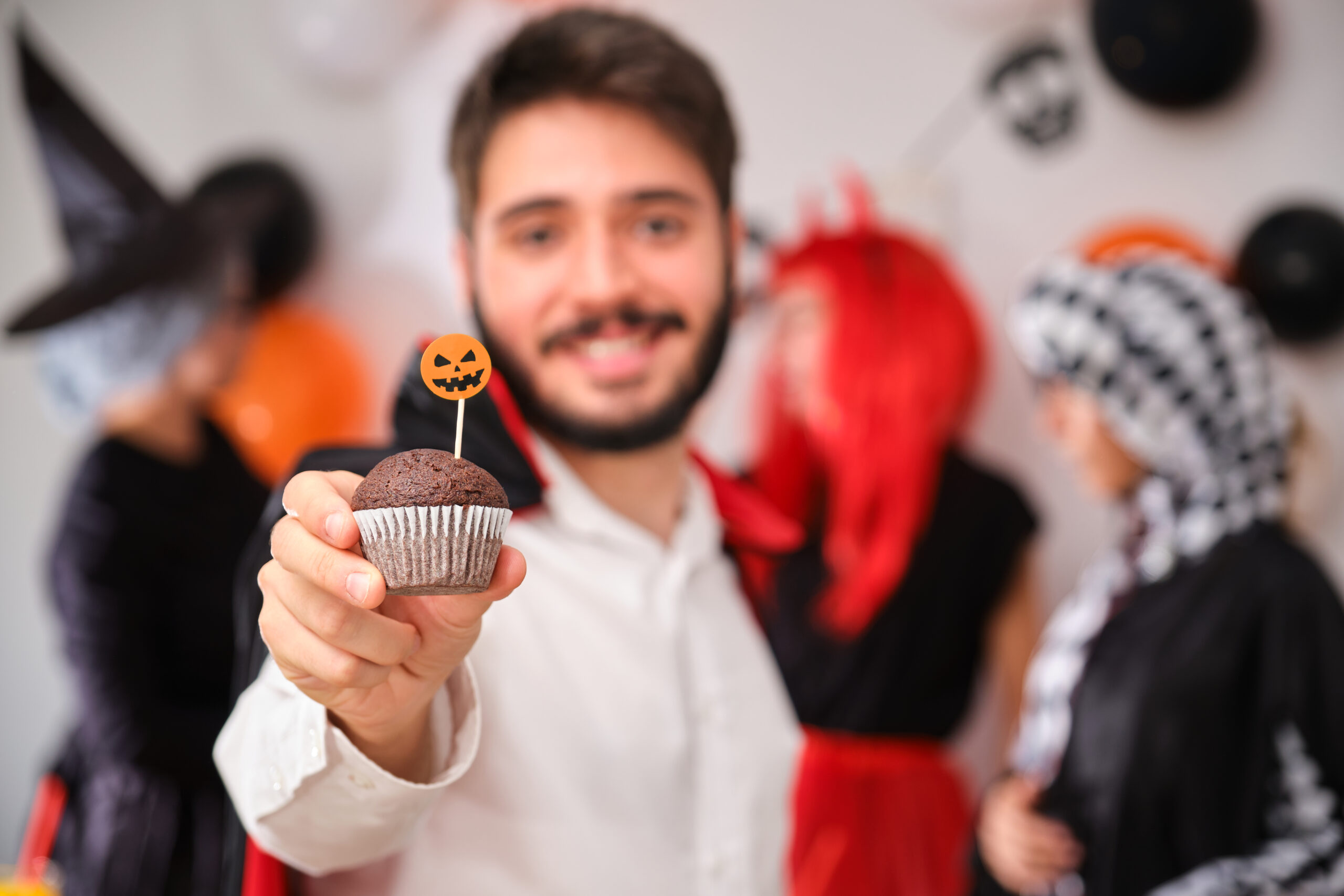 Young man in a Halloween costume holding a festive cupcake with a pumpkin topper, with coworkers in costumes celebrating in the background.