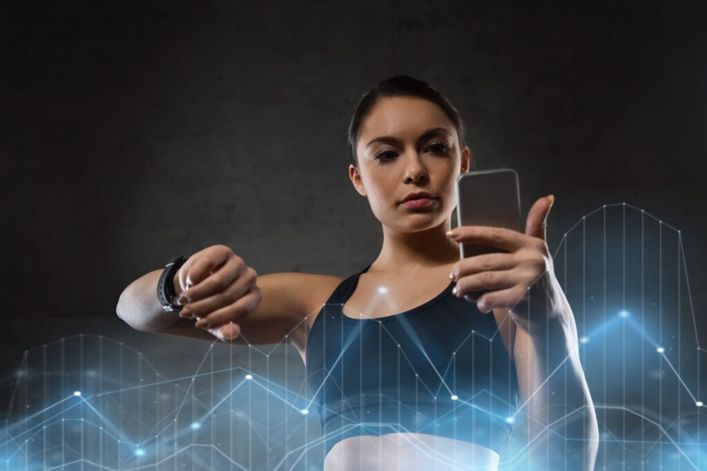A woman wearing a fitness tracker looks at her smartwatch and smartphone, analyzing health and wellness data displayed through digital graphs.