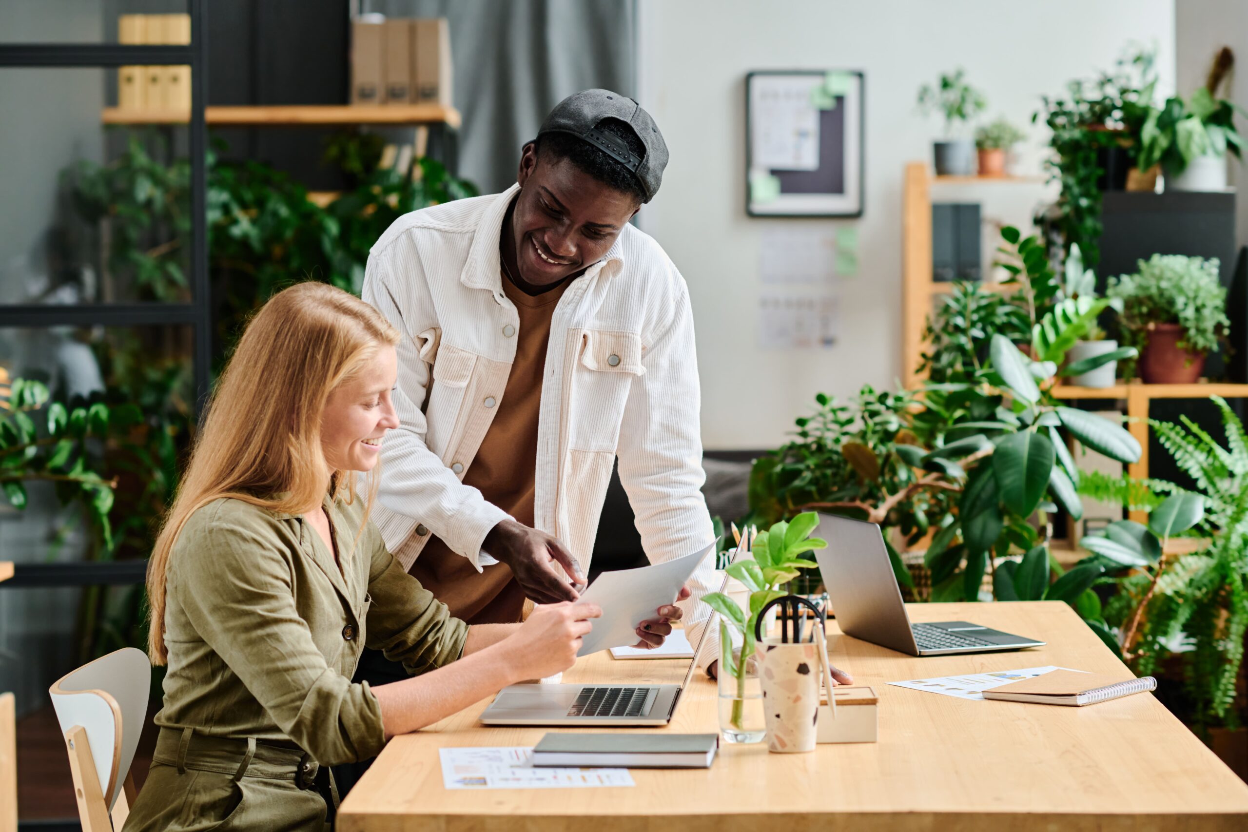 A founder and team member collaborate at a desk, smiling while reviewing documents in a bright, plant-filled workspace, symbolizing renewed motivation and positive energy.