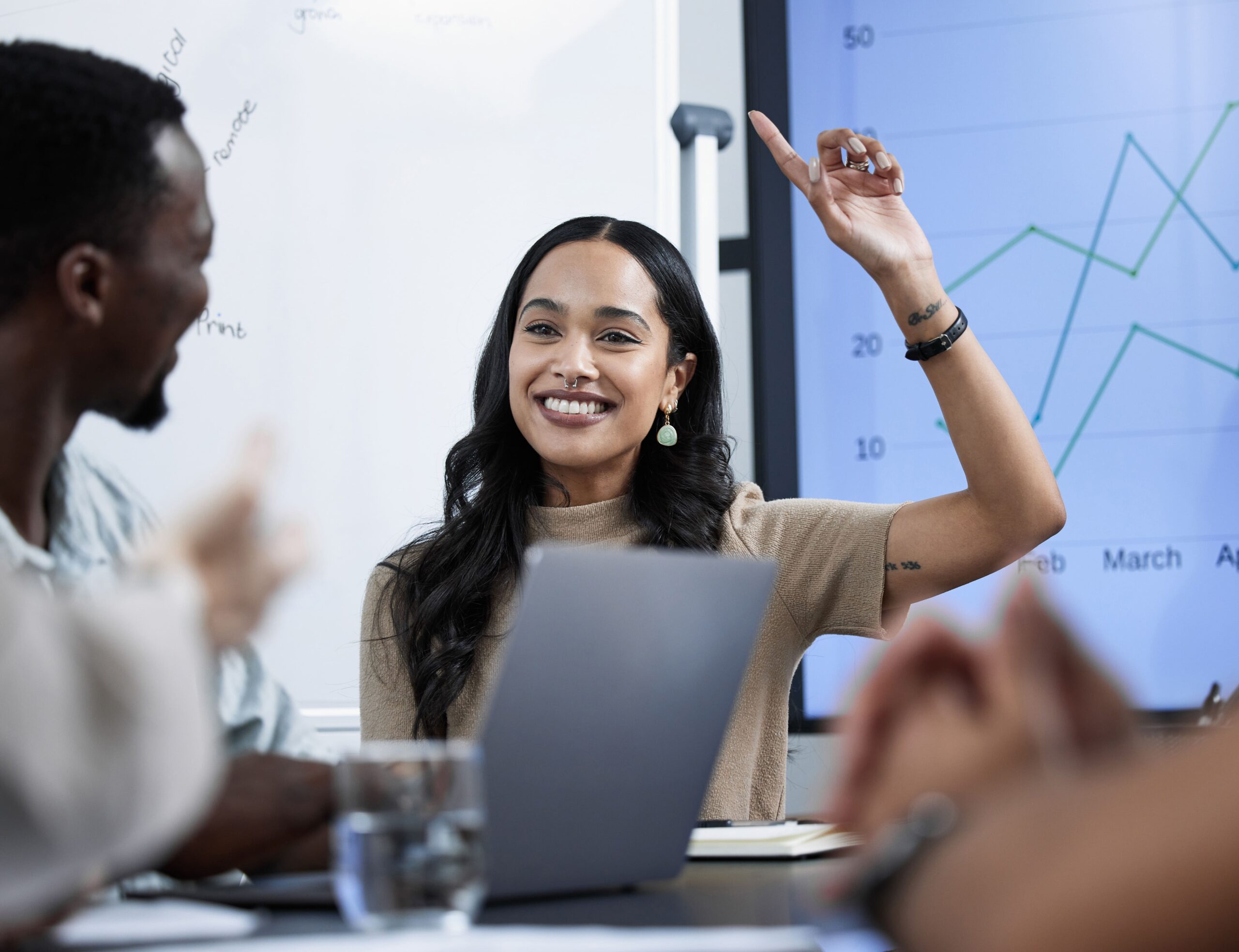 A smiling woman raises her hand during a team meeting, sitting at a table with coworkers and a laptop, with a line graph displayed on a screen behind her.