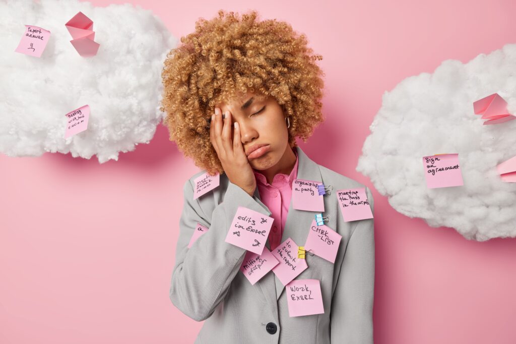 A stressed young woman in a suit stands against a pink background with her hand on her face, looking overwhelmed, covered in sticky notes listing tasks, symbolizing burnout and overload.