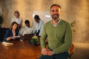 Confident business leader smiling while seated at a conference table, with a diverse team collaborating in the background — representing leadership resilience and team wellbeing in a startup environment.