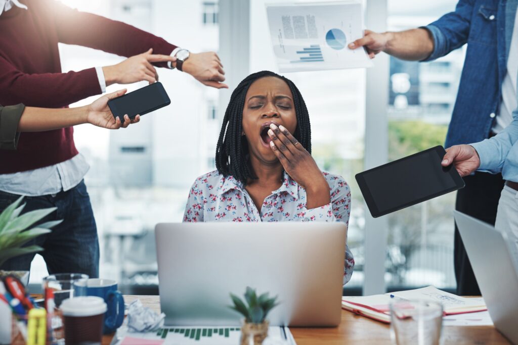 Stressed startup founder yawning at her desk surrounded by coworkers demanding attention with charts, devices, and time pressure — symbolizing burnout and the toll of constant demands.