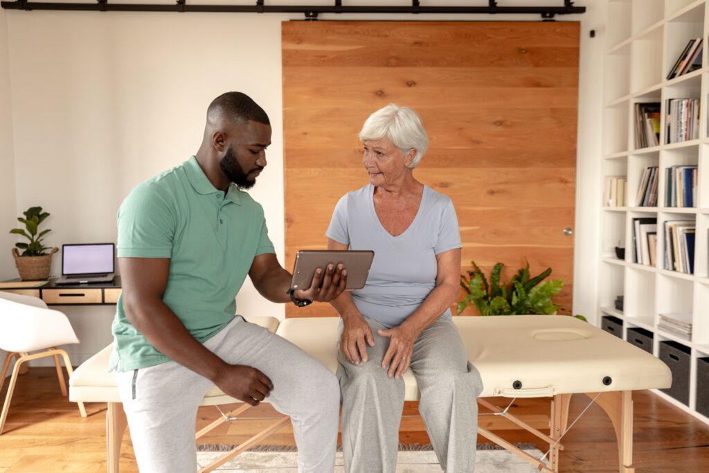 Physical therapist showing a personalized wellness plan on a digital tablet to an older woman during a consultation in a bright, modern clinic.