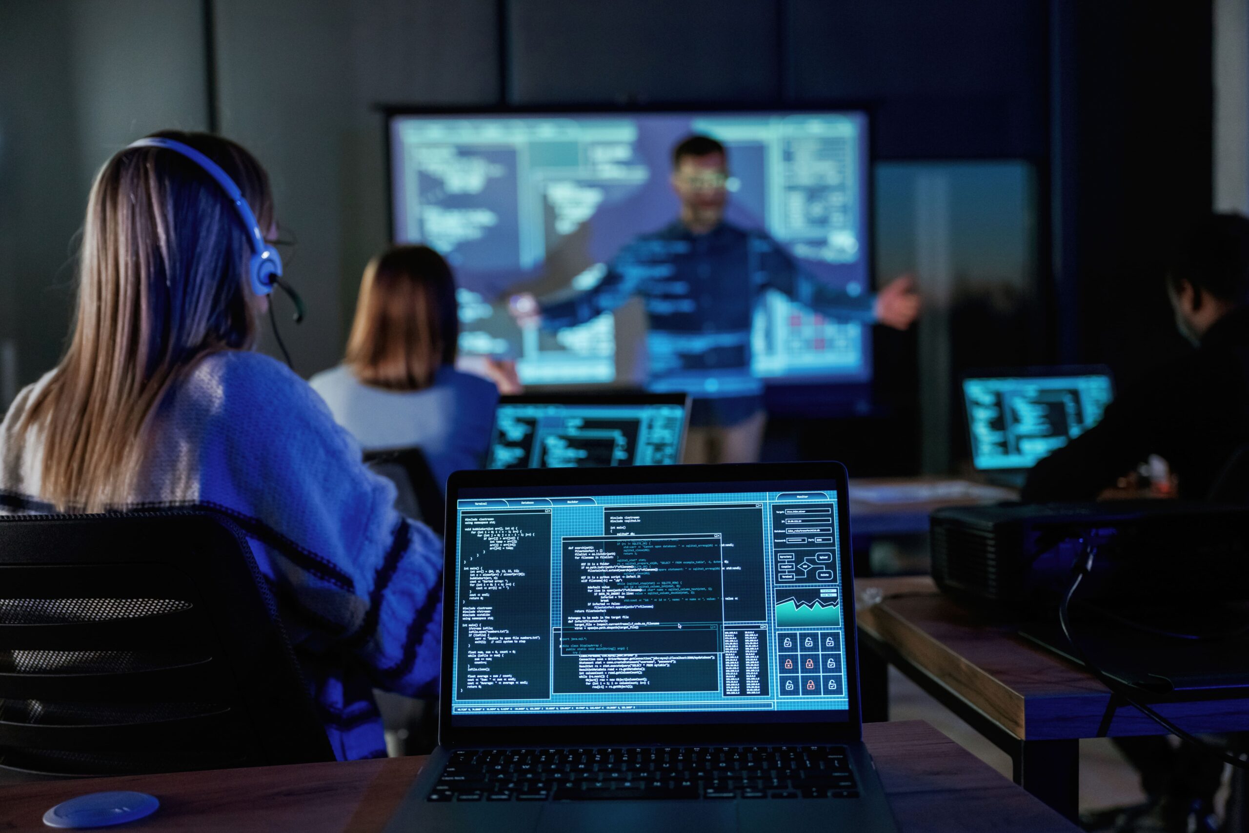 Employees participate in a cybersecurity training session, with laptops showing code and a presenter explaining threats on a projected screen.