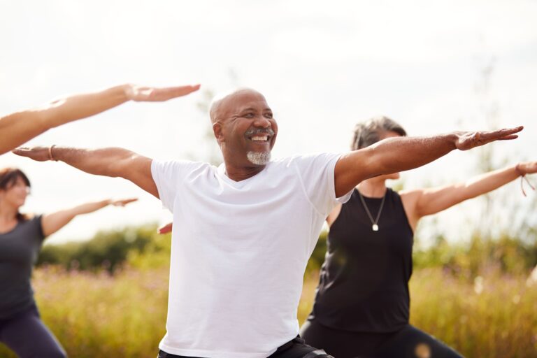 A group of adults participating in an outdoor yoga class, with a smiling man in the foreground practicing a stretching pose, representing wellness, movement, and preventative health programs for employers and TPAs.