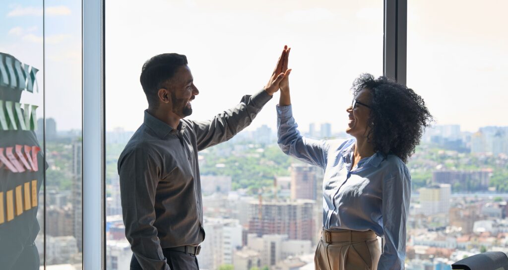 Two business professionals smiling and giving a high-five in a bright office with a city skyline in the background, symbolizing successful collaboration and achieving meaningful outcomes.