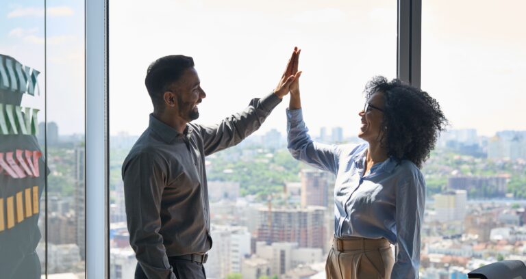 Two business professionals smiling and giving a high-five in a bright office with a city skyline in the background, symbolizing successful collaboration and achieving meaningful outcomes.
