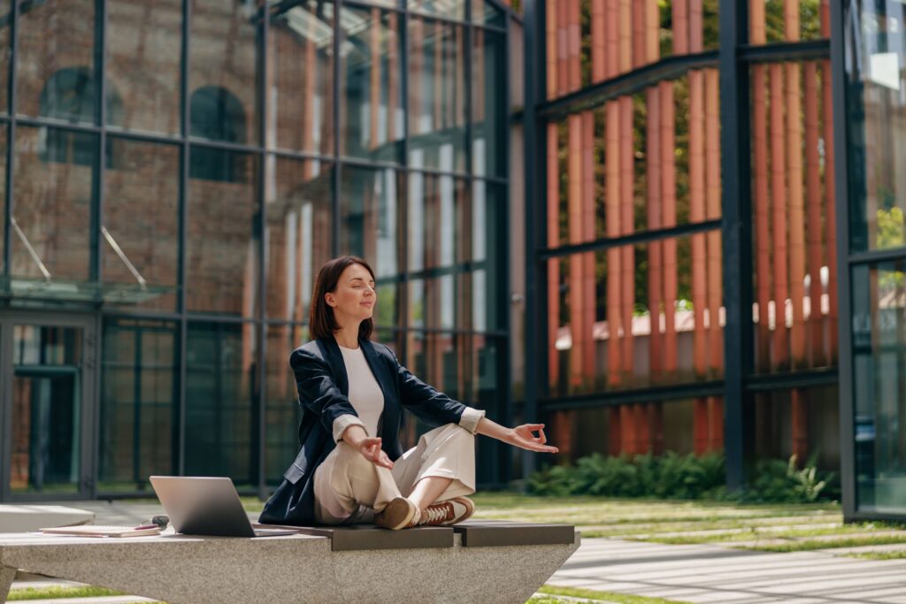 A professional woman sits outdoors in a meditation pose with her laptop beside her, symbolizing wellbeing and prevention as essential components of modern TPA value-added services.