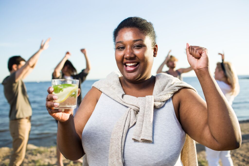 A smiling woman enjoys a drink at a beach gathering while friends celebrate in the background, symbolizing wellbeing and healthier lifestyles that help reduce diabetes-related claims.