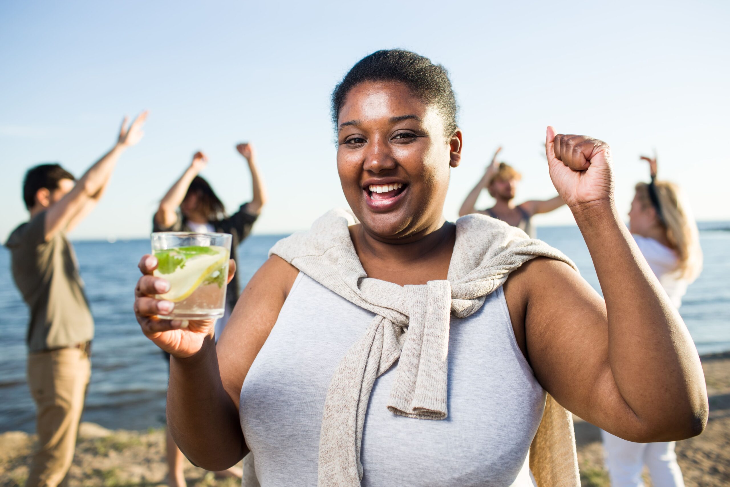 A smiling woman enjoys a drink at a beach gathering while friends celebrate in the background, symbolizing wellbeing and healthier lifestyles that help reduce diabetes-related claims.