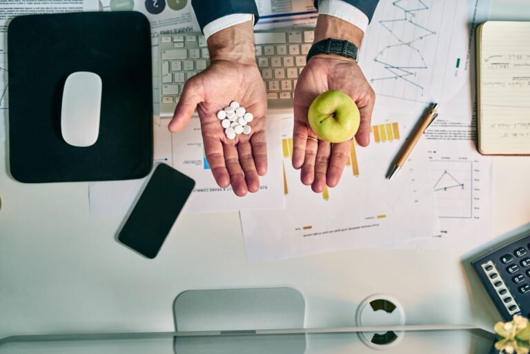 A business professional sits at a desk holding white medication pills in one hand and a green apple in the other, symbolizing the choice between medical treatment and preventive wellness amid healthcare data and reports.