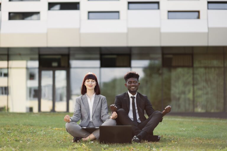 Two business professionals sit outdoors in a meditation pose, eyes closed and smiling, demonstrating workplace wellbeing and stress-reduction practices that support TPA employer retention strategies.