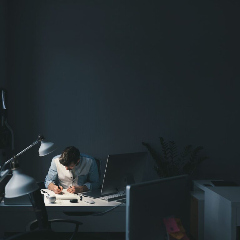 An employee working alone late at a desk under dim lighting, focused on tasks, illustrating quiet workplace burnout, sustained workload pressure, and unseen stress.