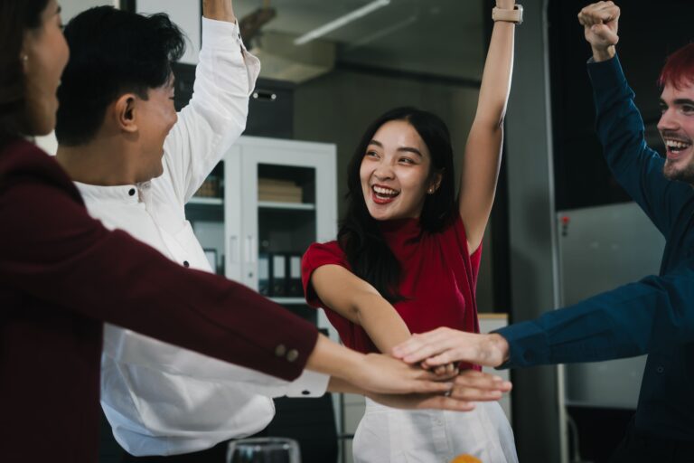 A diverse group of coworkers celebrating together in an office, stacking hands and raising arms to represent teamwork, recognition, and a strong workplace culture.