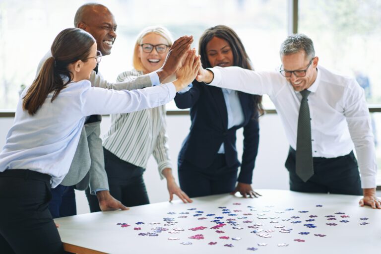 Business team celebrating progress together around a table, representing outcomes-based wellbeing, collaboration, and sustainable performance at work.
