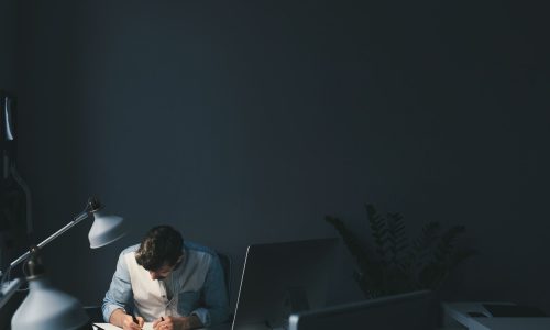 An employee working alone late at a desk under dim lighting, focused on tasks, illustrating quiet workplace burnout, sustained workload pressure, and unseen stress.