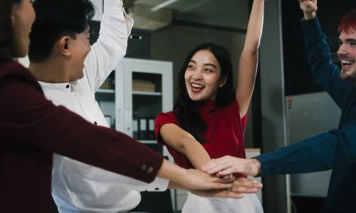 A diverse group of coworkers celebrating together in an office, stacking hands and raising arms to represent teamwork, recognition, and a strong workplace culture.