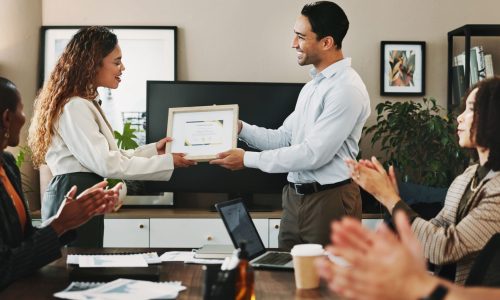 Manager presents a framed employee recognition award during a team meeting as colleagues applaud, highlighting appreciation, belonging, and recognition embedded in workplace culture.