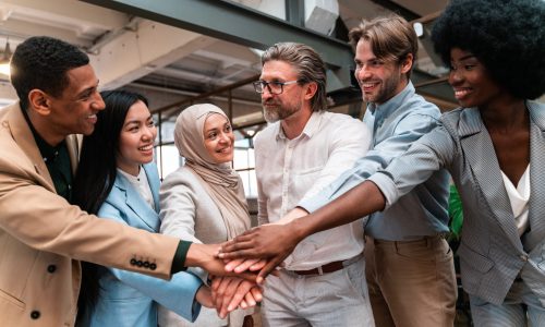 Diverse group of employees standing in a modern office with hands stacked together in the center, symbolizing teamwork, belonging, and a welcoming employee onboarding experience.